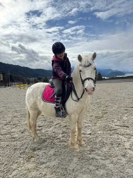 Ein Kind mit Reithelm sitzt auf einem weißen Pony unter weitem Wolkenhimmel.