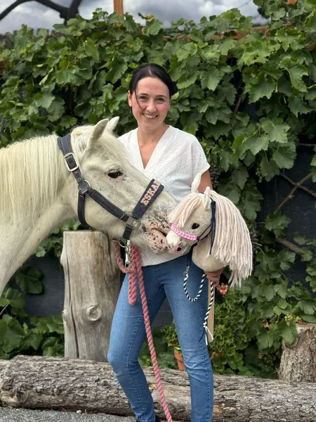 Eine Frau steht mit einem weißen Pony und einem Steckenpferd vor grünem Laub.