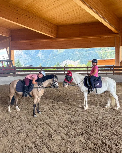Zwei Kinder sitzen auf Ponys in der offenen Reithalle mit Bergblick.