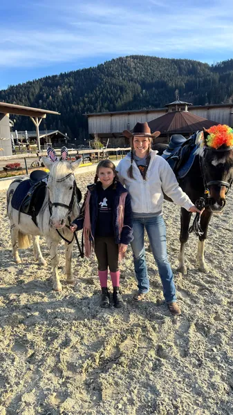 Ein Kind und eine Frau stehen zwischen zwei geschmückten Ponys auf dem Reitplatz.