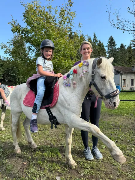 Ein Kind mit Reithelm sitzt auf einem weißen Pony neben einer Begleitperson auf der Wiese.