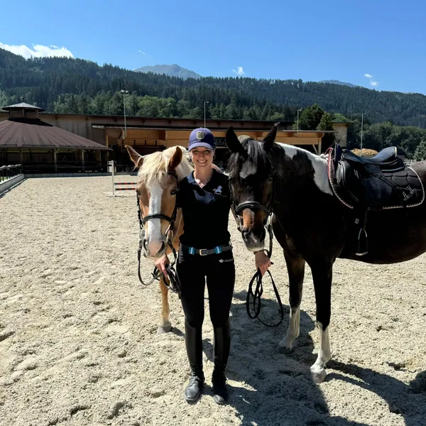 Eine Frau steht zwischen zwei gesattelten Ponys auf dem Reitplatz.