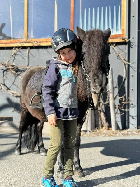 Ein Kind mit Reithelm steht eng neben einem dunklen Pony in der Sonne.